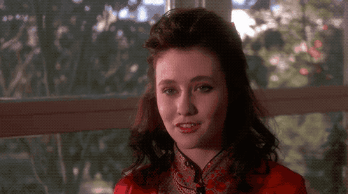 A Woman In A Red Jacket Is Smiling In Front Of A Window With Trees In The Background GIF