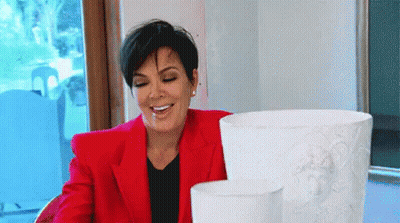 A Woman In A Red Jacket Is Smiling While Sitting At A Table With Two Vases . GIF