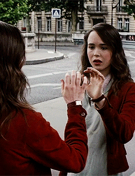 A Woman In A Red Jacket Talks To Another Woman In Front Of A Crosswalk Sign That Says A GIF