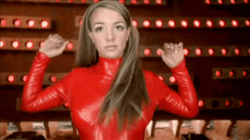 A Woman In A Red Leather Outfit Is Standing In Front Of A Wall Of Lights And Flexing Her Muscles . GIF