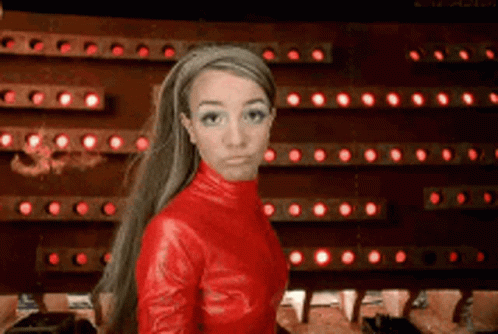 A Woman In A Red Leather Outfit Is Standing In Front Of A Wall Of Red Lights GIF