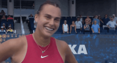 A Woman In A Red Nike Tank Top Smiles In Front Of A Ka Sign GIF