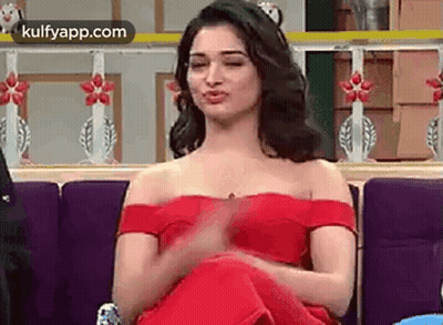 A Woman In A Red Off The Shoulder Dress Is Sitting On A Couch Making A Funny Face . GIF
