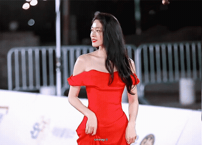 A Woman In A Red Off The Shoulder Dress Stands On A White Carpet GIF
