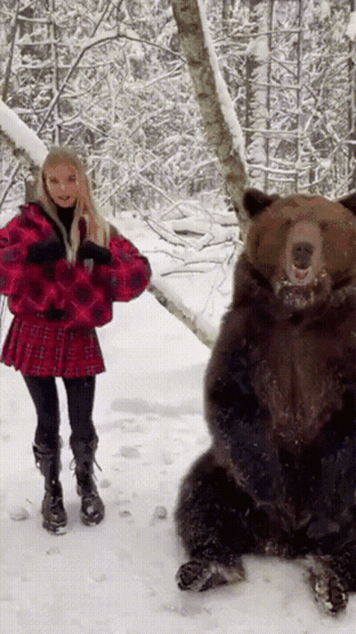A Woman In A Red Plaid Skirt Stands Next To A Bear In The Snow GIF