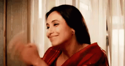 A Woman In A Red Saree Is Smiling And Clapping Her Hands In Front Of A Window . GIF