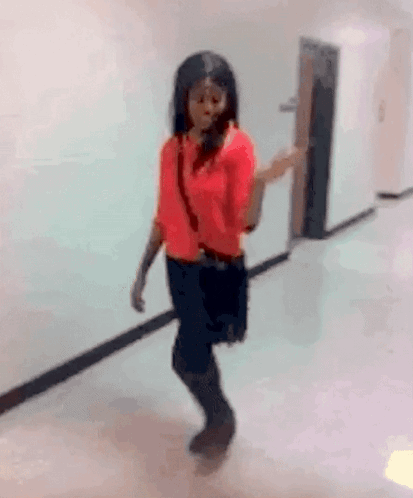 A Woman In A Red Shirt And Black Skirt Is Walking Down A Hallway GIF