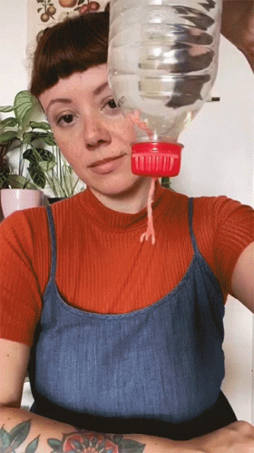 A Woman In A Red Shirt And Blue Tank Top Is Holding A Plastic Bottle With A Measuring Cup On It GIF