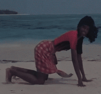 A Woman In A Red Shirt And Plaid Skirt Is Kneeling On A Beach GIF