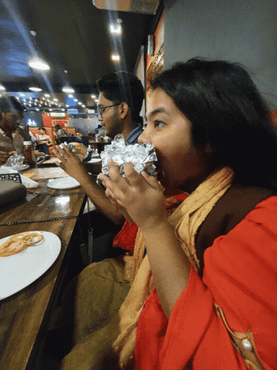 A Woman In A Red Shirt Is Eating A Wrapped Sandwich In A Restaurant GIF