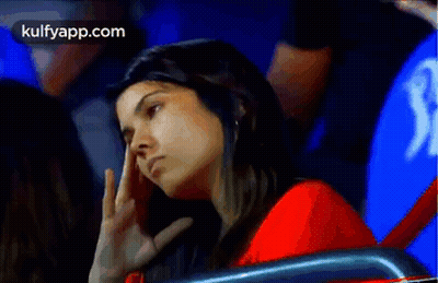 A Woman In A Red Shirt Is Sitting In A Stadium With Her Hand On Her Forehead . GIF