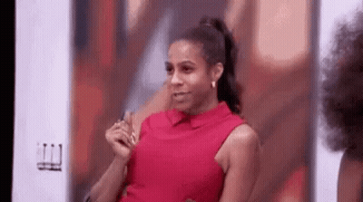A Woman In A Red Shirt Is Sitting In Front Of A Wall And Making A Funny Face . GIF