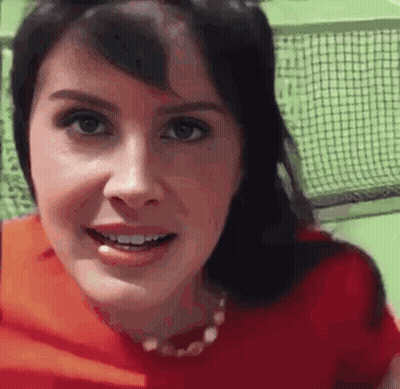 A Woman In A Red Shirt Is Smiling And Looking At The Camera While Standing In Front Of A Tennis Net . GIF