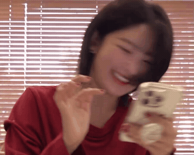 A Woman In A Red Shirt Is Smiling While Holding A Cell Phone In Front Of A Window With Blinds . GIF