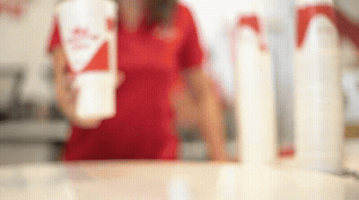 A Woman In A Red Shirt Is Standing Behind A Counter Holding A Cup Of Milkshake . GIF