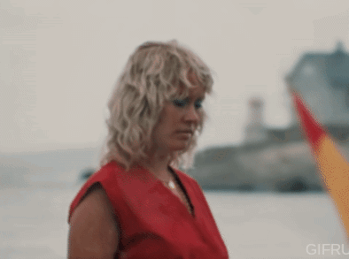 A Woman In A Red Shirt Is Standing In Front Of A Body Of Water GIF