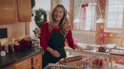 A Woman In A Red Sweater And Green Apron Is Standing In A Kitchen Preparing Christmas Cookies . GIF