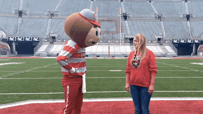 A Woman In A Red Sweatshirt Talks To A Mascot On A Field GIF