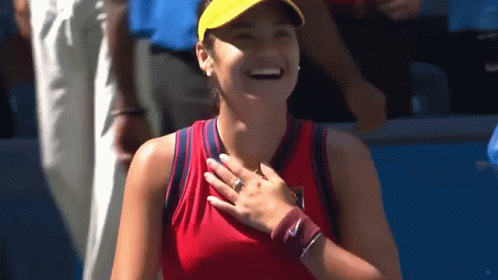 A Woman In A Red Tank Top And Yellow Hat Is Smiling And Holding Her Hand To Her Chest . GIF