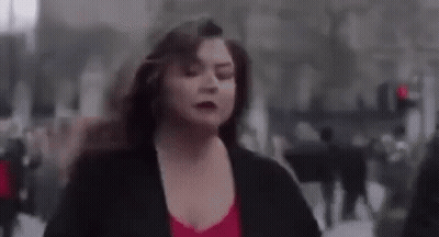 A Woman In A Red Top And Black Jacket Is Running Down A City Street . GIF