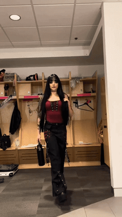 A Woman In A Red Top And Black Pants Is Standing In A Locker Room Holding A Water Bottle GIF