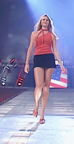 A Woman In A Red Top And Black Skirt Is Walking Down A Stage GIF