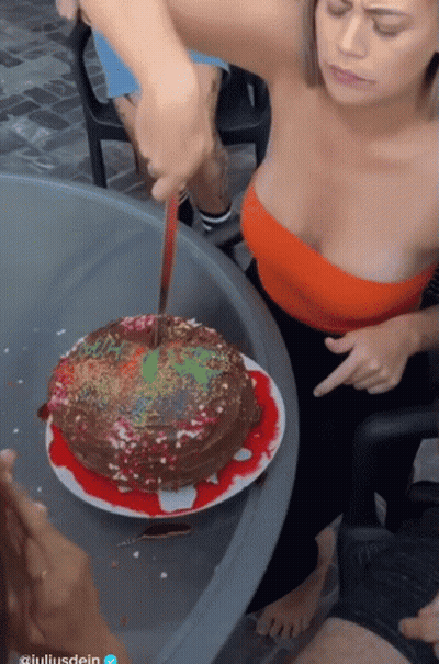 A Woman In A Red Top Is Cutting A Cake With A Knife In Front Of A Sign That Says Giuliardein GIF