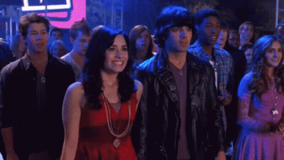 A Woman In A Red Top Stands Next To A Man In A Leather Jacket With Studs On It GIF