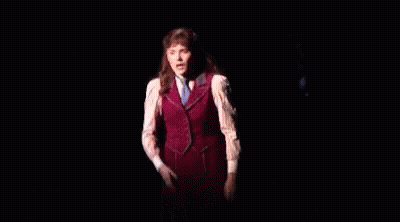 A Woman In A Red Vest And White Shirt Is Standing On A Stage In The Dark GIF