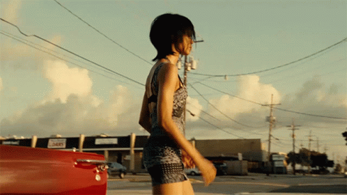 A Woman In A Short Dress Is Walking Down A Street In Front Of A Building That Says ' 365 ' On It GIF