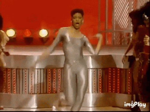 A Woman In A Silver Bodysuit Is Dancing On A Stage . GIF