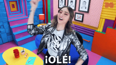 A Woman In A Silver Jacket Is Sitting In A Colorful Room With Her Arms In The Air And Says " Ole " GIF