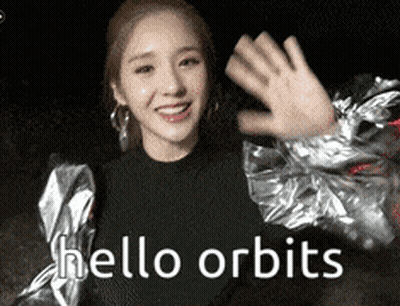 A Woman In A Silver Jacket Is Waving Her Hand And Says Hello Orbits GIF