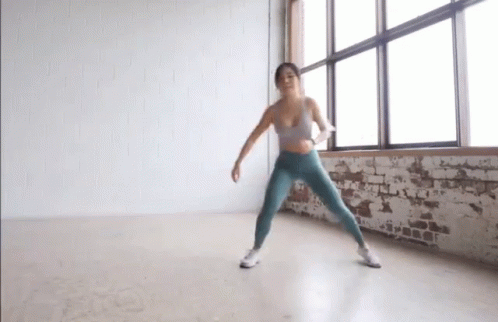 A Woman In A Sports Bra And Leggings Is Dancing In A Room In Front Of A Window . GIF