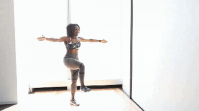 A Woman In A Sports Bra And Leggings Is Standing On One Leg In Front Of A Window With Her Arms In The Air GIF
