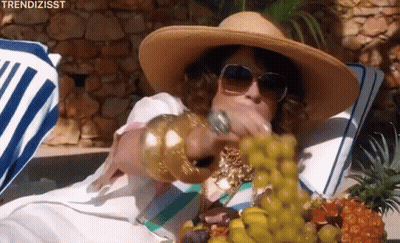A Woman In A Straw Hat And Sunglasses Is Eating Grapes While Laying On A Beach Towel . GIF