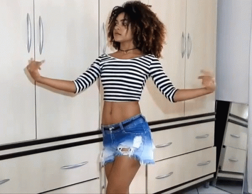 A Woman In A Striped Crop Top And Shorts Stands In Front Of A Dresser GIF