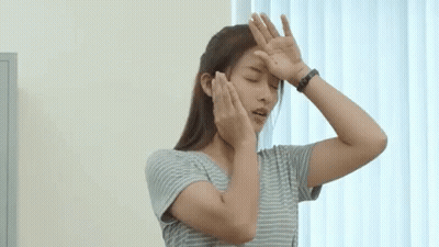 A Woman In A Striped Shirt Is Covering Her Face With Her Hands While Standing In Front Of A Window . GIF