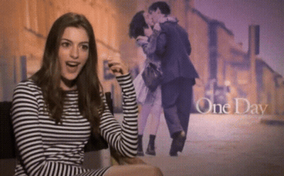 A Woman In A Striped Shirt Is Sitting In Front Of A Picture Of A Man And Woman Kissing With The Words One Day Above Them GIF