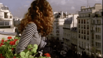 A Woman In A Striped Shirt Is Standing On A Balcony Looking Out Over A City GIF