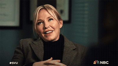 A Woman In A Suit And Black Turtleneck Is Smiling With The Nbc Logo Behind Her GIF