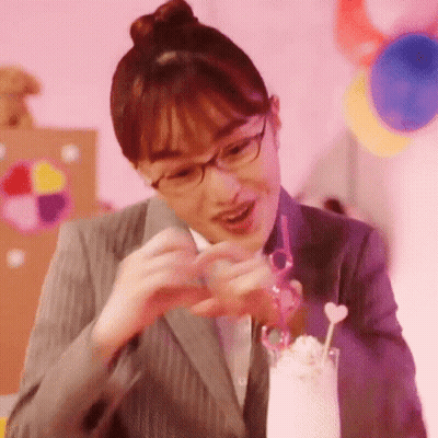 A Woman In A Suit And Glasses Is Making A Heart With Her Hands Next To A Glass Of Milkshake . GIF