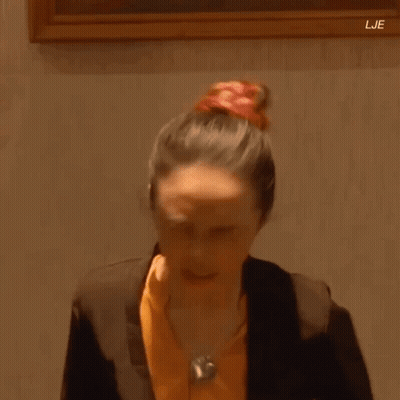 A Woman In A Suit And Orange Shirt Is Standing In Front Of A Framed Painting . GIF