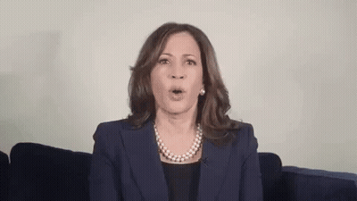 A Woman In A Suit And Pearl Necklace Is Sitting On A Couch With Her Mouth Open . GIF
