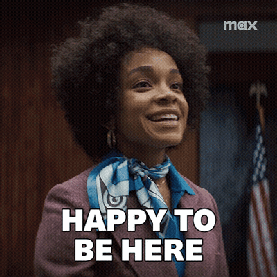 A Woman In A Suit And Scarf Is Smiling And Says " Happy To Be Here " GIF