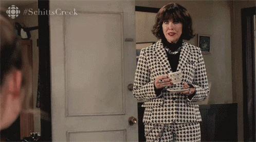 A Woman In A Suit Holds A Cup Of Coffee In Front Of A Door With #Schitts Creek Written On It GIF