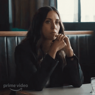 A Woman In A Suit Is Sitting At A Table With Her Hands Up In Front Of A Sign That Says Prime Video . GIF