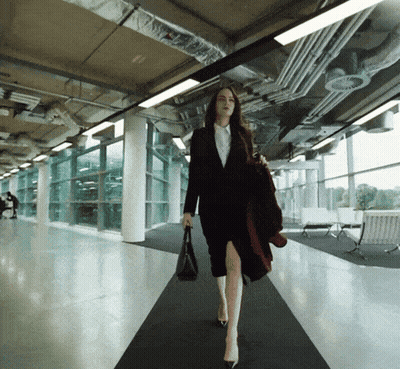 A Woman In A Suit Is Walking Down A Hallway Holding A Purse GIF