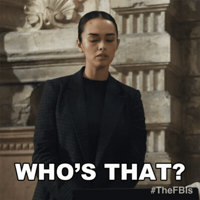 A Woman In A Suit Says " Who 'S That " In Front Of A Stone Wall GIF