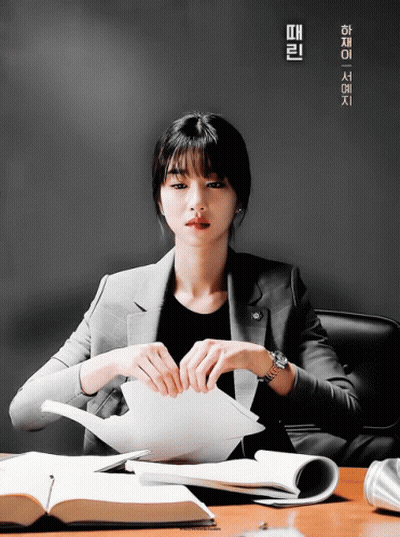 A Woman In A Suit Sits At A Desk Holding A Piece Of Paper In Front Of A Poster With Chinese Writing On It GIF